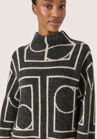 Mocha-mønstret sweater med høj hals, med et geometrisk design i cremefarvet på en mørkegrå baggrund, lavet af blødt uldlignende materiale.