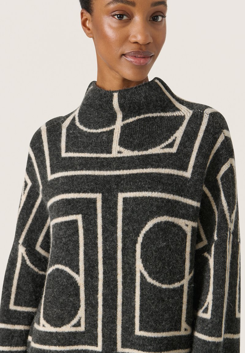 Mokka-musterter Pullover mit hohem Kragen, der ein geometrisches Design in Creme auf einem dunkelgrauen Hintergrund zeigt, aus weichem, wollartigem Material gefertigt.