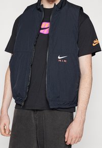 Mörkblå ärmlös väst med en texturerad yta, med en framzipper, tv å fickor och Nike-logotyp i vitt och rosa. Matchas med en svart t-shirt.