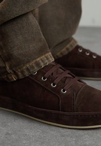 Baskets en daim marron foncé avec bout rond, œillets en métal et design de lacets texturé. Accentué par une semelle en caoutchouc de couleur claire.