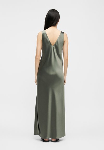 Femeie cu păr lung și închis la culoare, purtând o rochie din satin verde măsliniu, fără mâneci, lungă până în podea, cu o spate adânc în V, stând pe un fundal alb simplu.