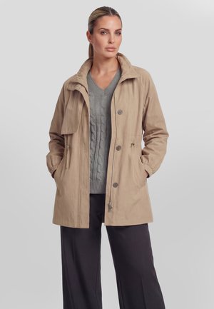 Donna in piedi con le mani in tasca, indossa una giacca beige abbottonata, un maglione grigio a trecce e pantaloni neri, rivolta in avanti.