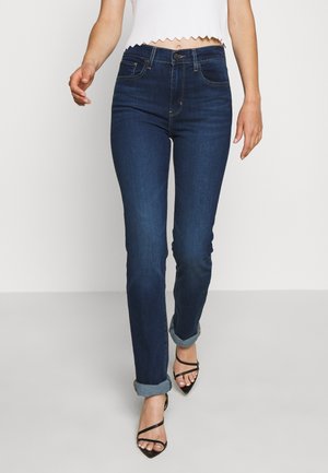 Femme portant un jean skinny bleu foncé retroussé et des sandales à talons hauts noires à brides, marchant devant un fond blanc uni.