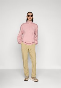 Felpa zip-up rosa chiaro, pantaloni beige e sneaker beige. Modella con occhiali da sole, in piedi su uno sfondo grigio uniforme.