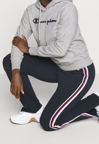 Person som knäböjer, iklädd en grå Champion-hoodie, marinblå träningsbyxor med vita och röda ränder, och vita sneakers mot en ljus bakgrund.