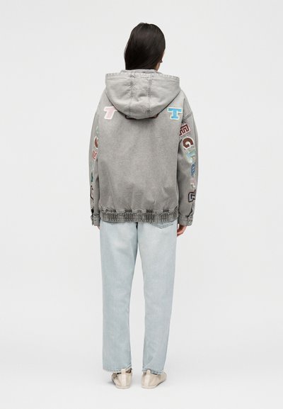 Άτομο που φορά γκρι μπλούζα με κουκούλα και πολύχρωμα γράμματα στα μανίκια και στην πλάτη, ανοιχτό μπλε τζιν και μπεζ αθλητικά παπούτσια, με την πλάτη γυρισμένη.