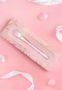 Różowo-złoty roller do twarzy z kamieniami szlachetnymi w przezroczystym pudełku z złotymi i różowymi wzorami geometrycznymi, na różowym tle.