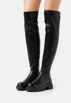 Bottines noires au-dessus du genou en cuir avec une texture lisse. Semelle en caoutchouc épaisse et détail de fermeture éclair sur le côté, avec une coupe ajustée.