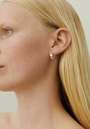 Szőke nő profilból, sima bőrrel, aki egy kicsi, fényes ezüst karika fülbevalót visel a bal fülében, semleges háttér előtt.