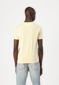 Tricou galben cu mânecă scurtă, confecționat dintr-un material moale, având un gât clasic și un accent în formă de V în spate, asortat cu blugi albaștri deschiși.
