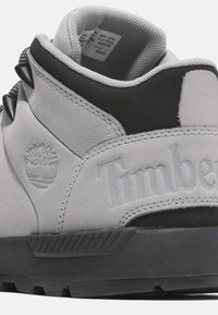 Grå mockaskor med svarta accenter, vadderad krage, texturerade snören och prägling av "Timberland"-logotypen på hälen. Gummi-sula med greppmönster.