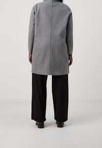Szary wełniany płaszcz o prostym kroju, długich rękawach i szwie z tyłu, zestawiony z czarnymi spodniami z szerokimi nogawkami oraz masywnymi czarnymi butami.