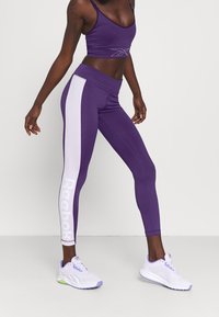 Lila träningsleggings med vita sidostråkar och "Reebok"-logotyp. Kombinerat med en matchande crop top och vit-lila sneakers.