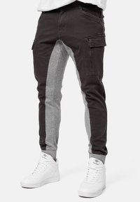 Cargo-Hosen mit einem Farbkombinationsdesign; dunkelbrauner und grauer Stoff, mit Seitentaschen und schmal zulaufenden Beinen. Kombiniert mit weißen Sneakern.