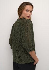 Olivenfarvet blondebluse med puffede ærmer, teksturelt blomstermønster, flæser ved halsen og en løs pasform, parret med mørke bukser.