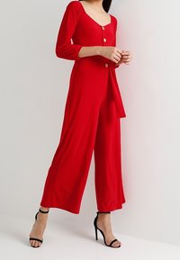 Mono rojo con mangas largas, escote en V profundo, botones en la parte delantera y detalle de cintura atada. Tela suave y silueta de pierna ancha. Sandalias de tacón negras.