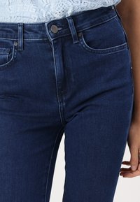 Pantalones vaqueros de mezclilla azul oscuro con un diseño ajustado, cintura alta y el clásico estilo de cinco bolsillos. Textura suave con sutiles detalles de costura.
