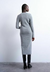 Femme aux cheveux tressés portant une robe midi gris ajustée en maille côtelée et des bottes noires montant jusqu'aux genoux, debout sur un sol en béton.