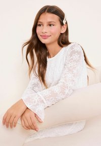 Valkoinen pitsipaita, jossa kukkakirjailu, pitkät hihat ja kellomaiset kalvosimet. Mallilla on pitkät ruskeat hiukset ja helmi hiuskoristeena. Tasainen tausta.