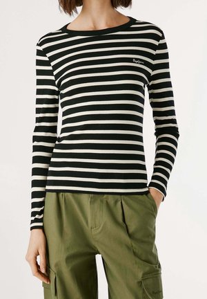 Femme portant un haut à manches longues rayé noir et blanc et un pantalon cargo vert olive, une main dans la poche, sur un fond uni.