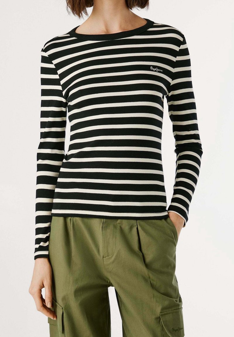 Femme portant un haut à manches longues rayé noir et blanc et un pantalon cargo vert olive, une main dans la poche, sur un fond uni.