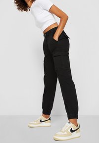 Pantaloni cargo neri con polsini elasticizzati, molteplici tasche e vestibilità rilassata, abbinati a sneaker beige e bianche con uno swoosh nero.