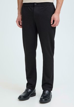 Mężczyzna ubrany w czarne spodnie o prostych nogawkach z elastycznym pasem oraz czarne skórzane buty wsuwane, stojący na jednolitym tle.