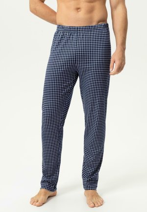 Man die marineblauwe en witte geruite pyjamabroek draagt, staat op blote voeten met één hand rustend op zijn heup.