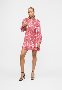 Νεαρή γυναίκα με πλεξούδες φοράει μακρυμάνικο φόρεμα με κόκκινο και λευκό μοτίβο, κρατά καφέ τσάντα ώμου και φορά μαύρα πέδιλα με ψηλό τακούνι.