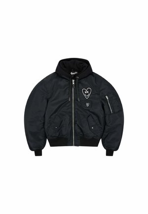 Jeune homme aux cheveux courts porte une veste bomber noire à capuche avec des patchs en forme de cœur, debout les mains jointes devant un fond uni.