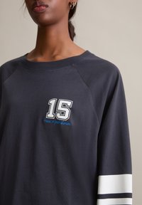 Donkergrijze sweatshirt met een losse pasvorm, voorzien van een witte "15" opdruk en blauwe tekst. Witte en marineblauwe strepen accentueren de mouwen.