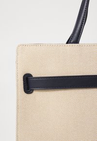 Beige geweven handtas met een textuur oppervlakte, voorzien van een gladde zwarte leren schouderband en details, rechthoekige vorm en minimalistisch ontwerp.