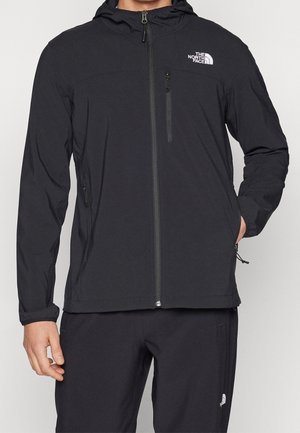 Man draagt een zwarte North Face-jas met capuchon, voorrits en met ritssluitingen op de borst- en zijzakken, gecombineerd met een zwarte broek.