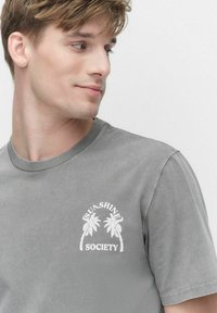 Grå T-shirt med korte ærmer lavet af blød bomuld, med hvid palmetræsgrafik og teksten "SUNSHINE SOCIETY" på brystet. Casual pasform.