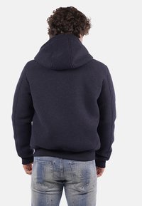 Felpa con cappuccio blu navy con finitura testurizzata. Presenta un cappuccio prominente e orlo a coste, abbinata a jeans chiari strappati.