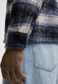 Wollene Karoschirt in Blau, Schwarz und Creme mit strukturierter Oberfläche, zu sehen mit einem geknöpften Manschette und Jeans.