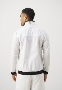 Fehér nylon bomber dzseki álló gallérral, fekete mandzsettákkal és derékkal, hátul vízszintes csíkkal. A textúrája sima.