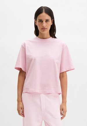 Kobieta o prostych ciemnych włosach w jasnoróżowej, oversize'owej koszulce i dopasowanych spodniach, stojąca na tle jednolitego białego tła.