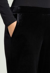 Zwarte fluwelen broek met een gladde textuur, voorzien van zijzakken en een zachte tailleband. Close-up weergave met nadruk op stof en designdetails.