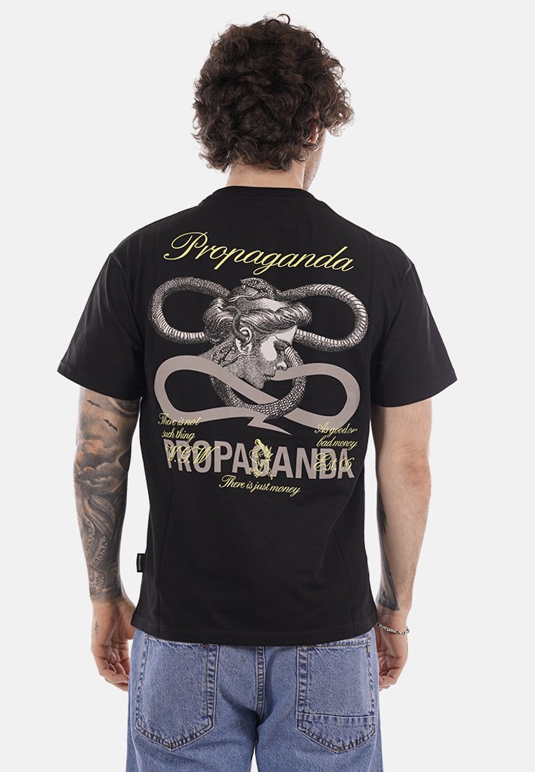 Uomo con capelli ricci che indossa una maglietta nera con un serpente che si avvolge intorno alla testa di una donna classica e il testo "Propaganda" e "C'è solo denaro."