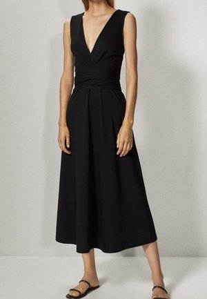 Woman wearing a sleeveless black midi dress with deep V-neck and black flat sandals, standing against a plain light background.