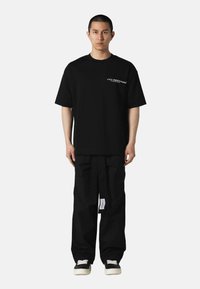 Svart bomulls oversized T-skjorte med hvit tekst, kombinert med løse svarte cargo-bukser med lommer og et etikettmerke. Svart sneakers fullfører antrekket.