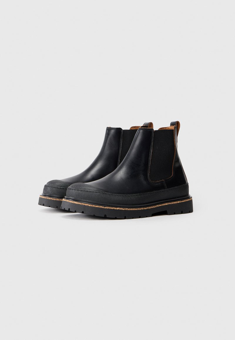 Une paire de bottines Chelsea en cuir noir avec des semelles en caoutchouc épaisses et des panneaux latéraux élastiques sur un fond gris clair.