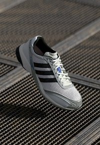 Hvid atletisk sko med mesh-overdel, sorte stribede detaljer, grå sål og forstærket tå. Snørebåndene er lys grå med et blåt detalje.