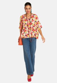 Floral-print, short-sleeve tunic in vibrant colors, paired with high-waisted jeans and red heels. Accessories include an orange bag and beaded necklace.