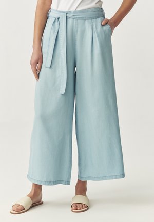Frau trägt hellblaue, weit geschnittene Hose mit Gürtel, weißes Oberteil und beige Slides-Sandalen, steht mit einer Hand in der Tasche.