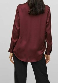 Burgundy satin blouse with long sleeves, buttoned cuffs, rounded hem, and a smooth texture, worn over black pants.