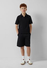 Zwarte polo met korte mouwen en een kraag, gecombineerd met bijpassende zwarte shorts. De outfit heeft een gestructureerd patroon en wordt aangevuld met witte sneakers.