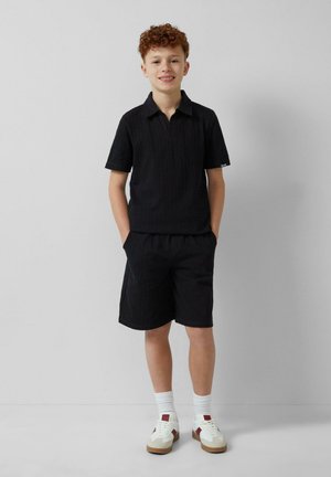 Zwarte polo met korte mouwen en een kraag, gecombineerd met bijpassende zwarte shorts. De outfit heeft een gestructureerd patroon en wordt aangevuld met witte sneakers.