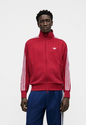 Muškarac u crvenoj Adidas jakni na patent s bijelim prugama na rukavima i plavim trenirkama na jednostavnoj bijeloj pozadini.